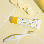 A 50g tube of Jack N' Jill banana flavoured fluoride free kid's toothpaste placed on a yellow tiled surface next to a toothbrush. 
