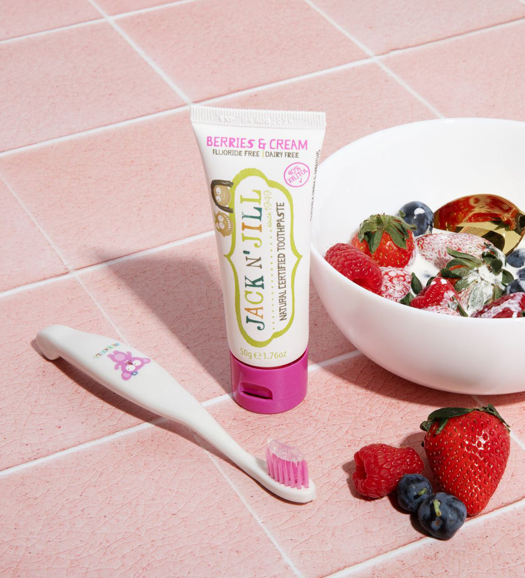 A 50g tube of Jack N' Jill berries and cream flavoured fluoride free kid's toothpaste placed on pink tiles next to a Jack N' Jill toothbrush. A bowl full of berries and cream can be seen to the side. 