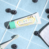 A 50g tube of Jack N' Jill blueberry flavoured fluoride free kid's toothpaste placed on a blue tiled surface next to a Jack N' Jill toothbrush surrounded by blueberries. 