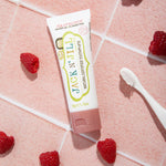 A 50g tube of Jack N' Jill raspberry flavoured fluoride free kid's toothpaste placed on a pink tiled surface surrounded by raspberries. 