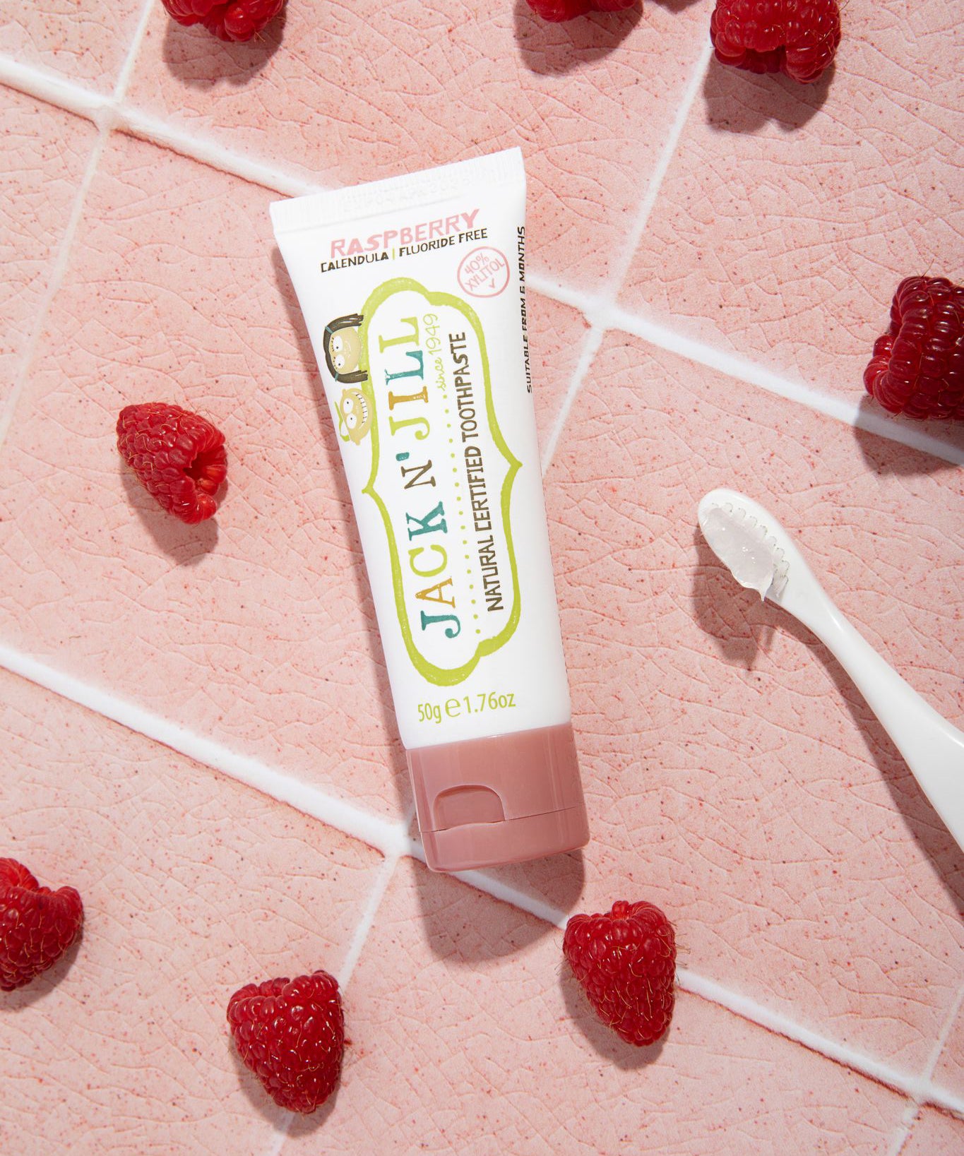 A 50g tube of Jack N' Jill raspberry flavoured fluoride free kid's toothpaste placed on a pink tiled surface surrounded by raspberries. 
