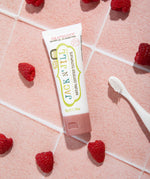 A 50g tube of Jack N' Jill raspberry flavoured fluoride free kid's toothpaste placed on a pink tiled surface surrounded by raspberries. 