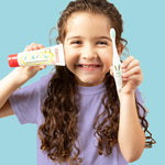 A child grinning and holding up a  50g tube of Jack N' Jill strawberry flavoured fluoride free kid's toothpaste next to a toothbrush. 