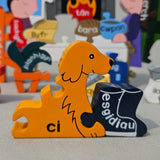 Jig-So Cymraeg Cartref. A Welsh Home themed wooden jigsaw puzzle with painted pieces.The photo shows a close up of the dog shaped piece with the Welsh for for dog ci written ion it. A selection of Welsh Jigsaw puzzles from Alphabet Jigsaws are available at Babipur.