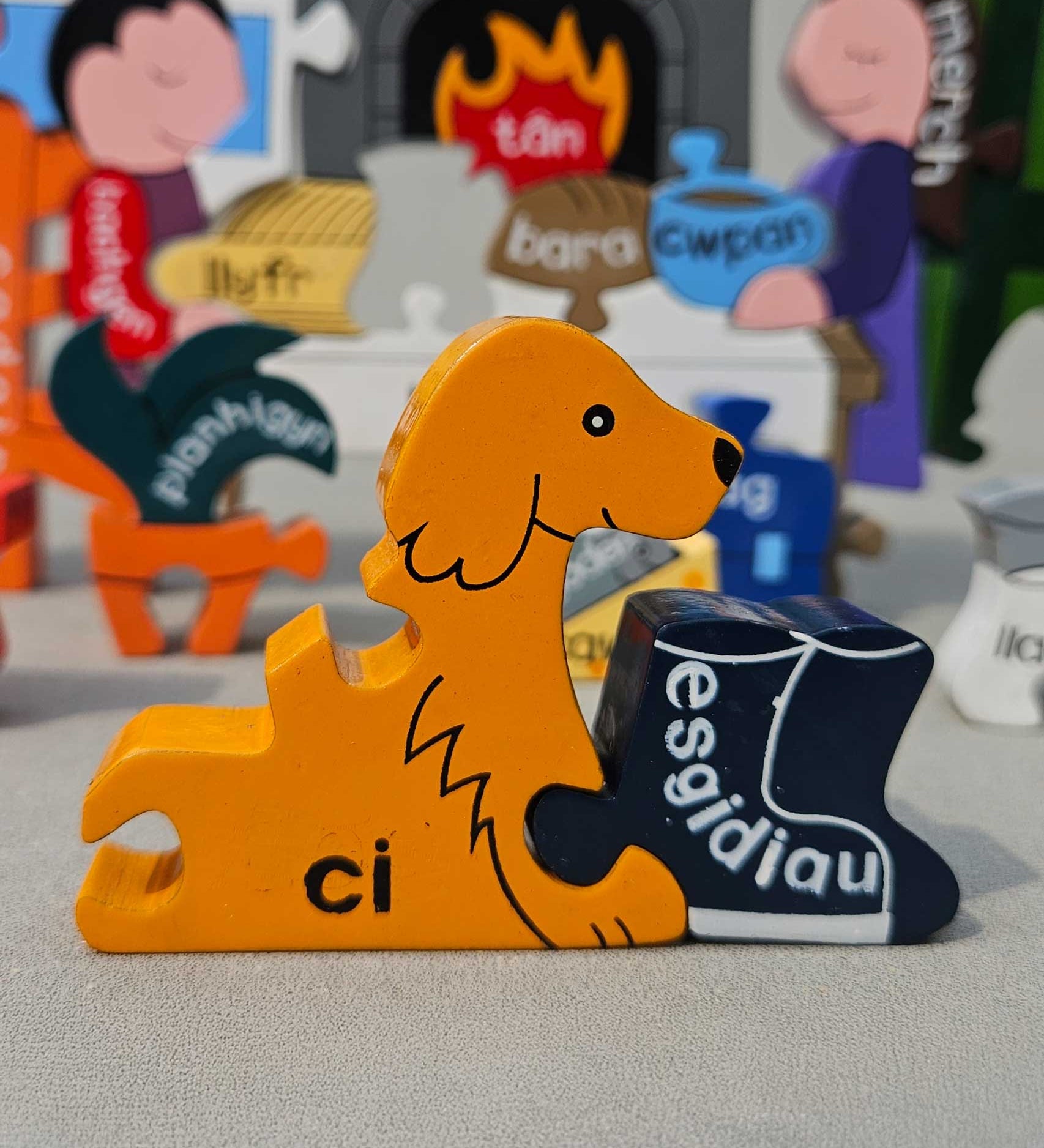 Jig-So Cymraeg Cartref. A Welsh Home themed wooden jigsaw puzzle with painted pieces.The photo shows a close up of the dog shaped piece with the Welsh for for dog ci written ion it. A selection of Welsh Jigsaw puzzles from Alphabet Jigsaws are available at Babipur.