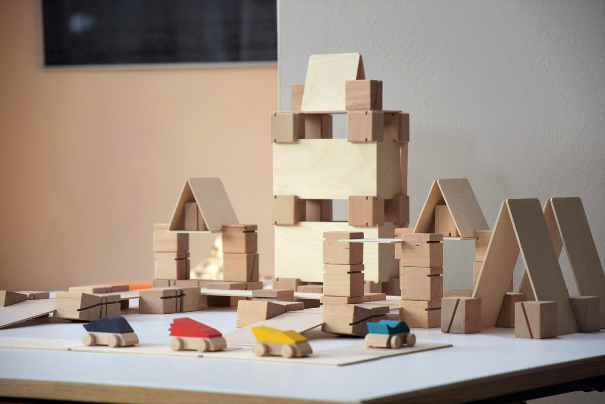 Just Blocks plastic-free wooden Smart Line Blocks stacked into the shape of a building behind some wooden toy cars