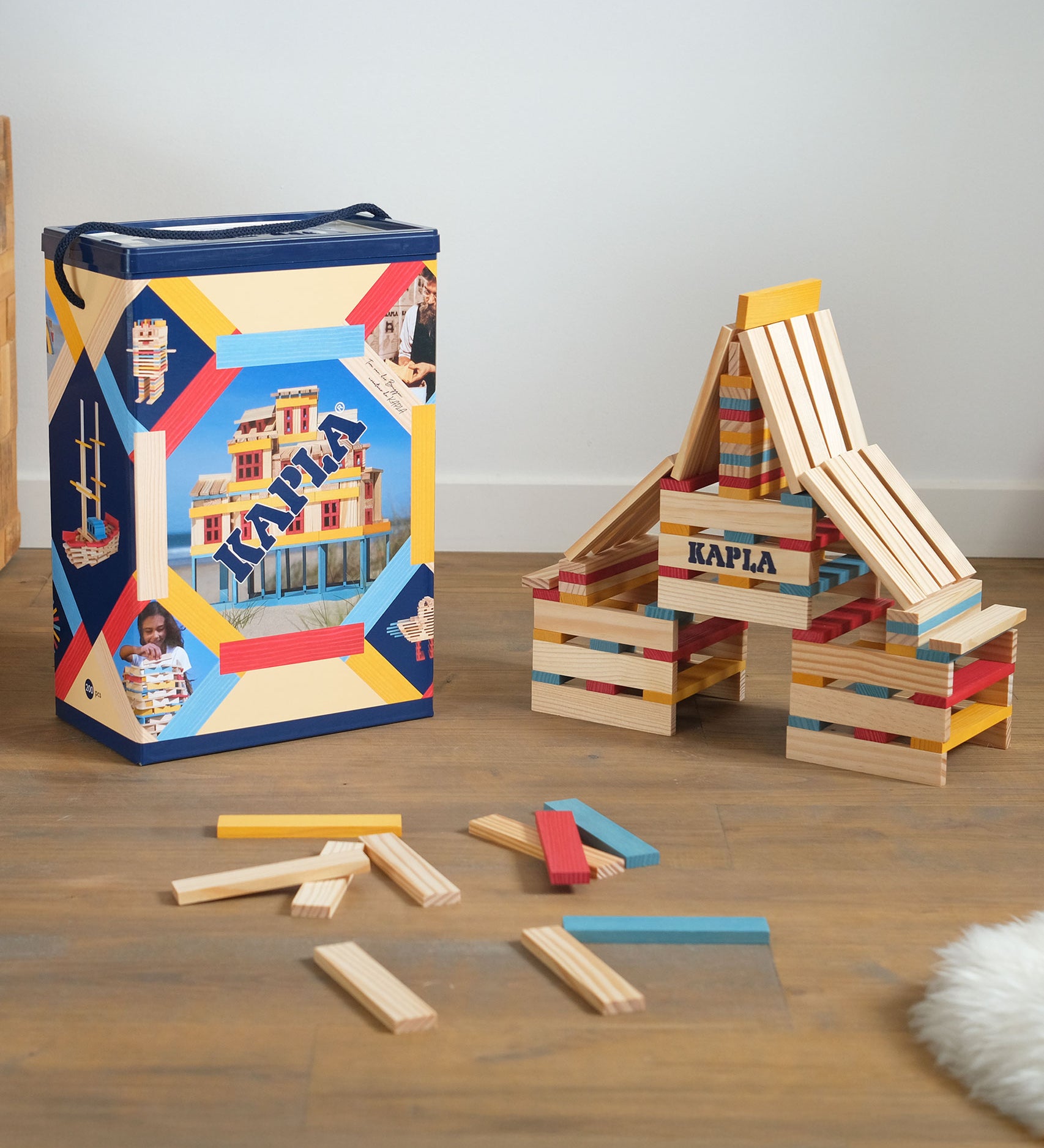 KAPLA Wooden Building Blocks 200 Box in Summer colours. A build can be seen by the side of the box with a few blocks on the floor. 