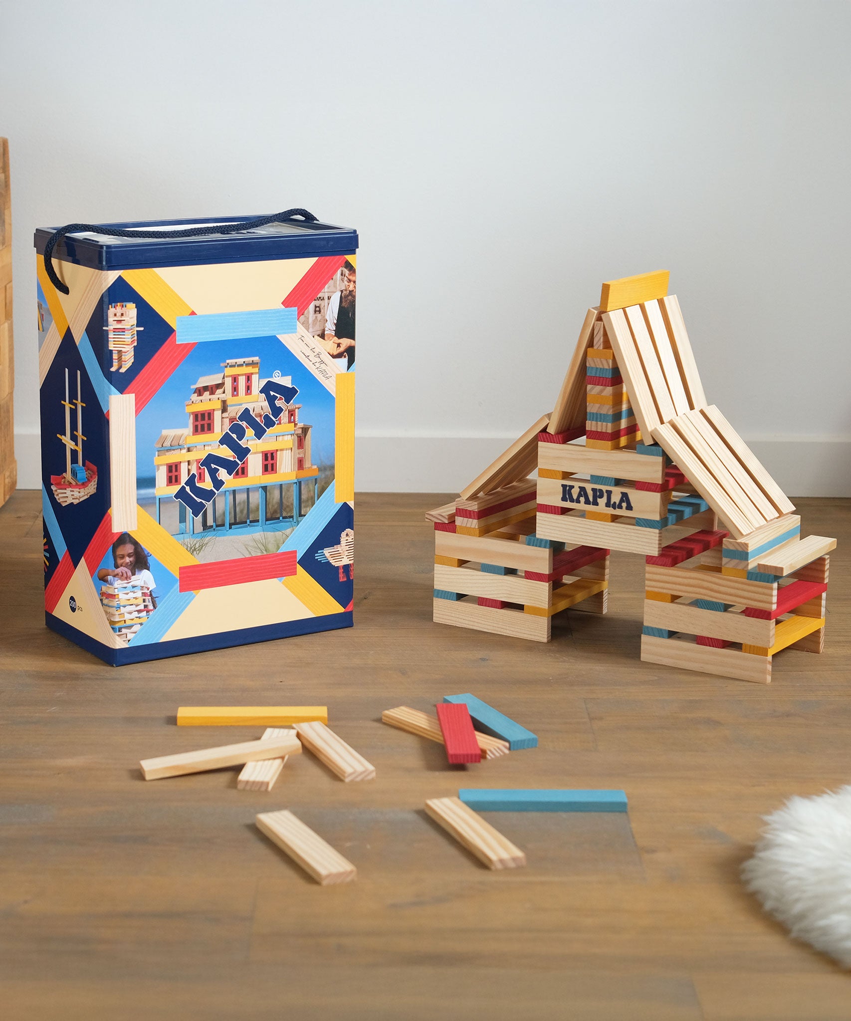 KAPLA Wooden Building Blocks 200 Box in Summer colours. A build can be seen by the side of the box with a few blocks on the floor. 
