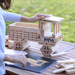 Child placing down a wooden Kapla building block on top of a model truck behind the advanced Kapla art book