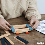 Close up of some hands stacking the Kapla natural and blue wooden toy blocks into a square shape on a wooden table