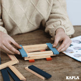 Close up of some hands stacking the Kapla natural and blue wooden toy blocks into a square shape on a wooden table