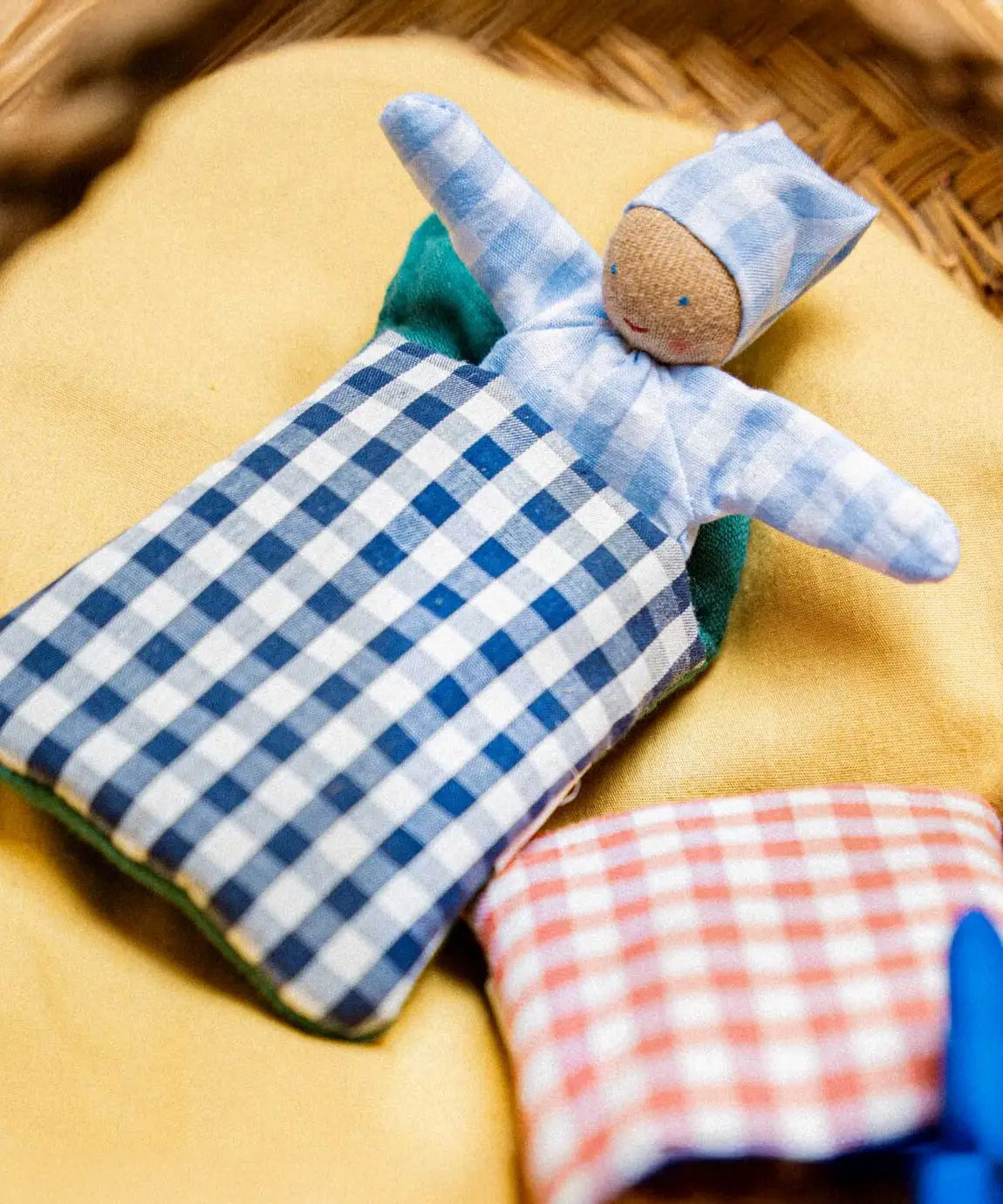 A blue waldorf kathe kruse doll inside a blue gingham blanket sitting inside a wicker basket
