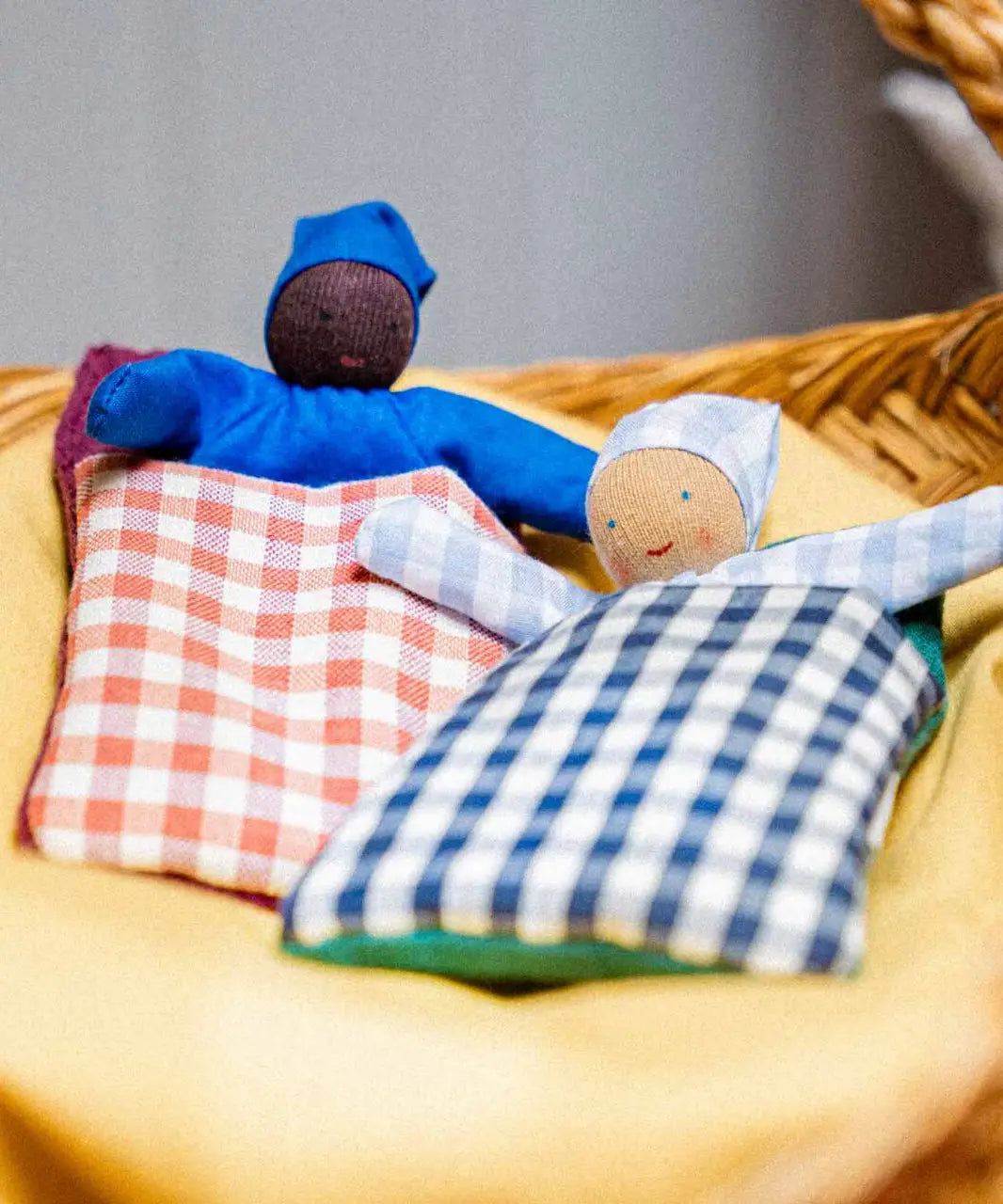 2 Kathe Kruse baby waldorf dolls lying inside gingham blankets on a wicker basket