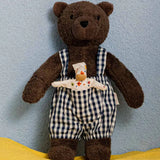 Käthe Kruse Bella brown bear soft toy wearing blue and white gingham dungaree standing on a yellow blanket