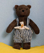 Käthe Kruse Bella brown bear soft toy wearing blue and white gingham dungaree standing on a yellow blanket