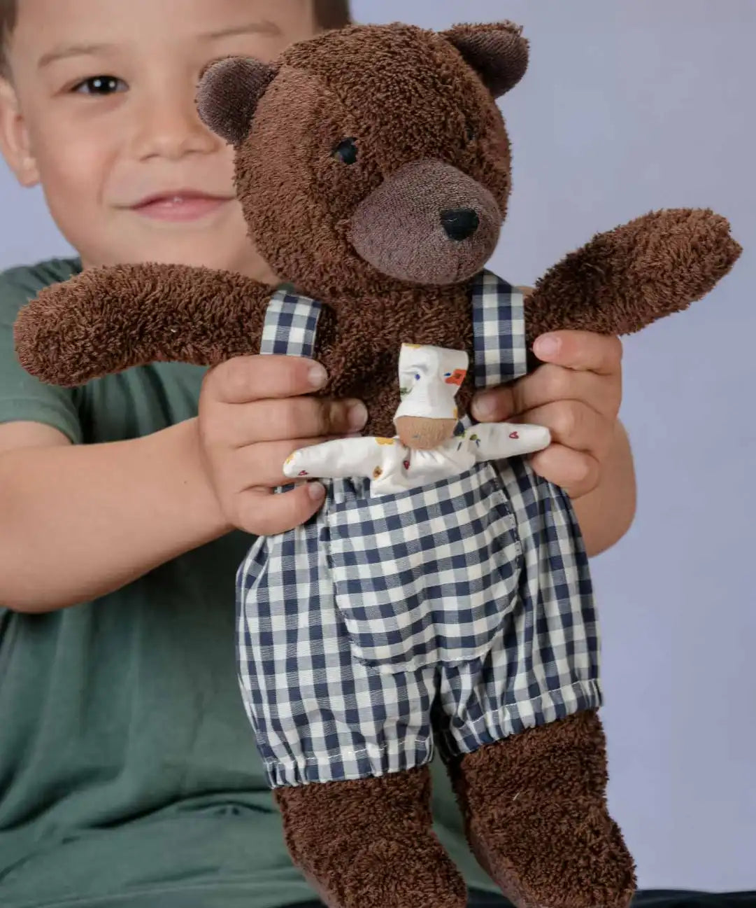 Käthe Kruse Bella brown bear soft toy wearing blue and white gingham dungarees with a small doll in it's front pocket.