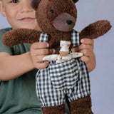 Käthe Kruse Bella brown bear soft toy wearing blue and white gingham dungarees with a small doll in it's front pocket.