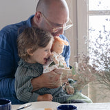 A child sitting in an adult's lap holding a Käthe Kruse Waldorf doll Elsa. Doll has a White patterned tunic top with pink linen style trousers underneath. White patterned bow in her hair with green and white sneaker style shoes.
