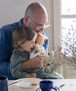 A child sitting in an adult's lap holding a Käthe Kruse Waldorf doll Elsa. Doll has a White patterned tunic top with pink linen style trousers underneath. White patterned bow in her hair with green and white sneaker style shoes.