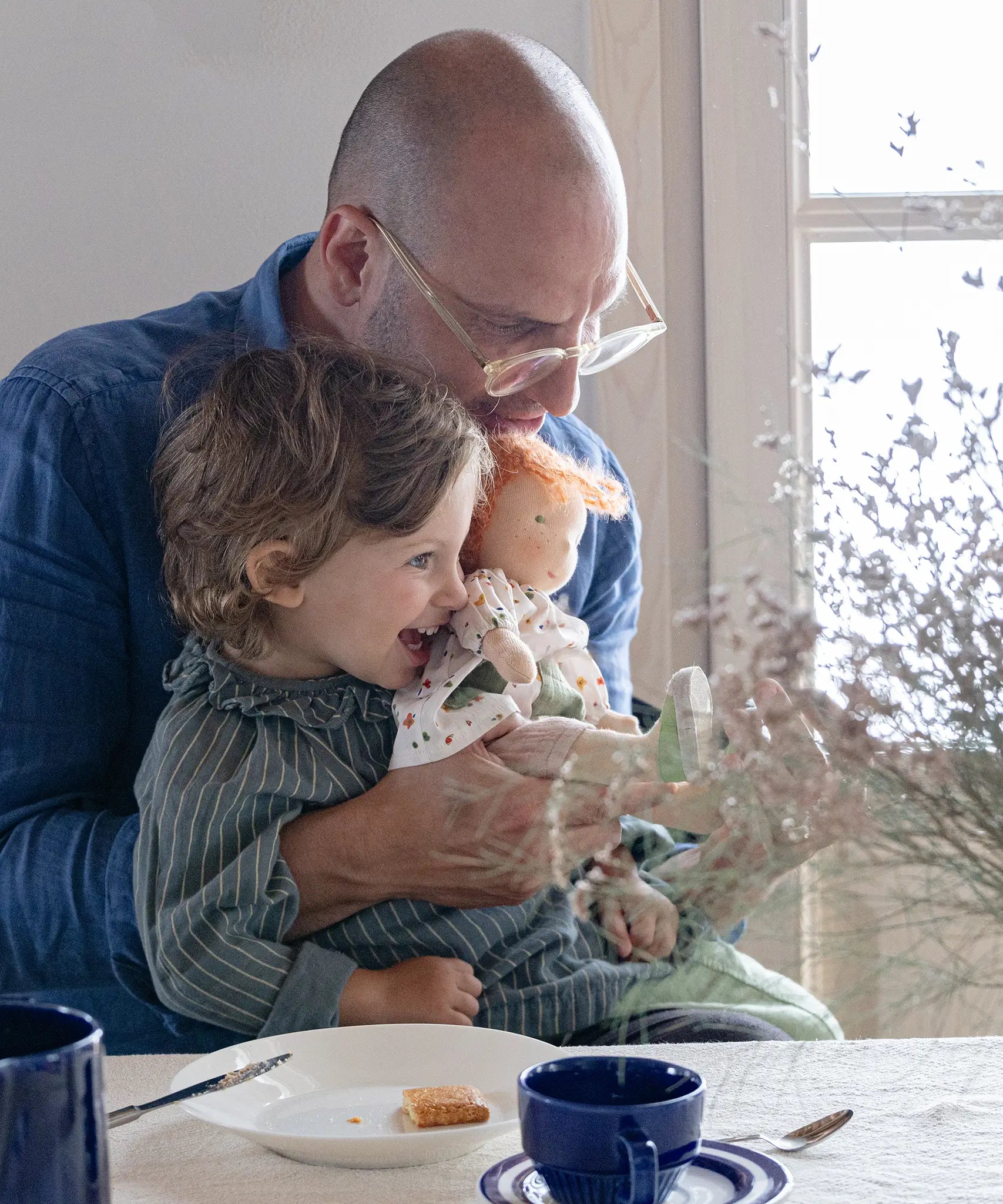 A child sitting in an adult's lap holding a Käthe Kruse Waldorf doll Elsa. Doll has a White patterned tunic top with pink linen style trousers underneath. White patterned bow in her hair with green and white sneaker style shoes.