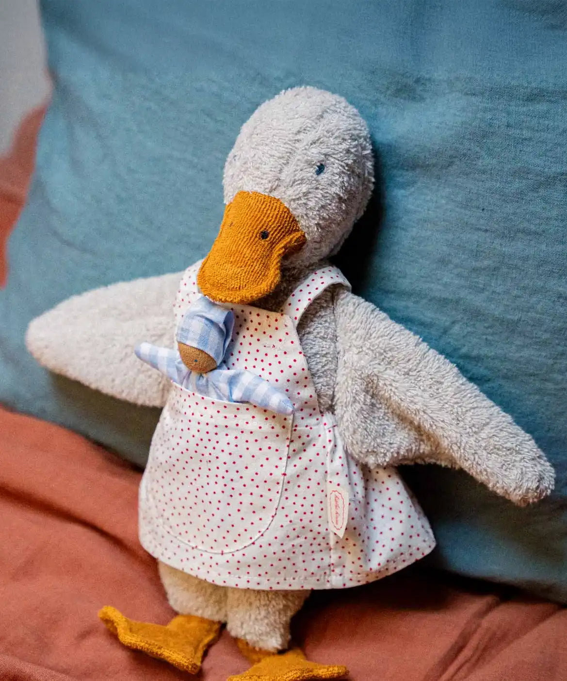 Käthe Kruse Eva duck standing soft toy wearing a white apron with red dots all over. There is a small doll in the apron pocket.