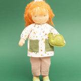 Käthe Kruse Waldorf doll Elsa with long red wavy hair and fair skin and a green felt snail. White patterned tunic top with pink linen style trousers underneath. White patterned bow in her hair with green and white sneaker style shoes.