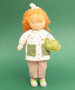 Käthe Kruse Waldorf doll Elsa with long red wavy hair and fair skin and a green felt snail. White patterned tunic top with pink linen style trousers underneath. White patterned bow in her hair with green and white sneaker style shoes.
