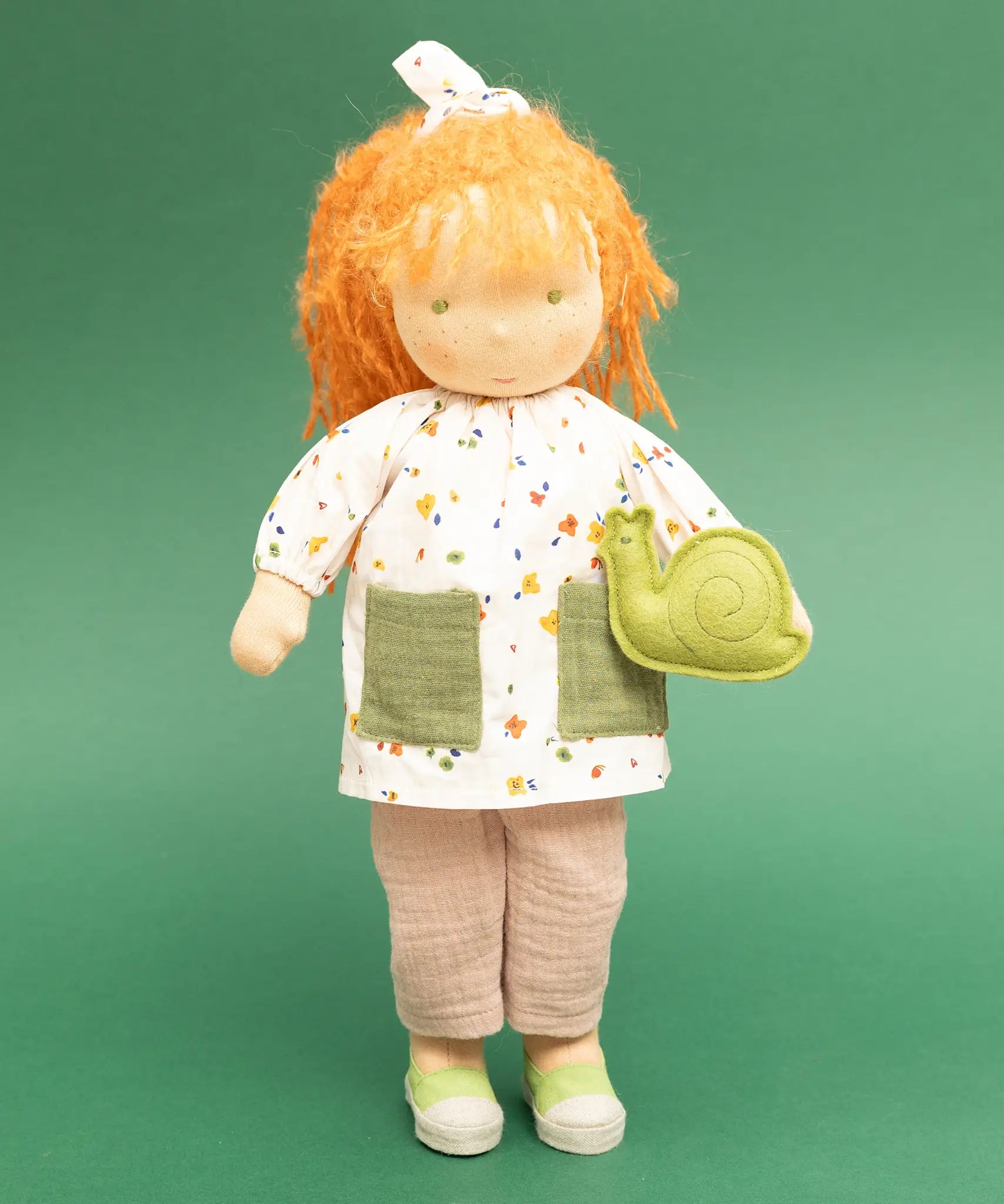 Käthe Kruse Waldorf doll Elsa with long red wavy hair and fair skin and a green felt snail. White patterned tunic top with pink linen style trousers underneath. White patterned bow in her hair with green and white sneaker style shoes.