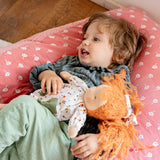 Child laying on a large pink pillow with aKäthe Kruse Waldorf doll Elsa with long red wavy hair and fair skin. White patterned tunic top with pink linen style trousers underneath. White patterned bow in her hair with green and white sneaker style shoes.