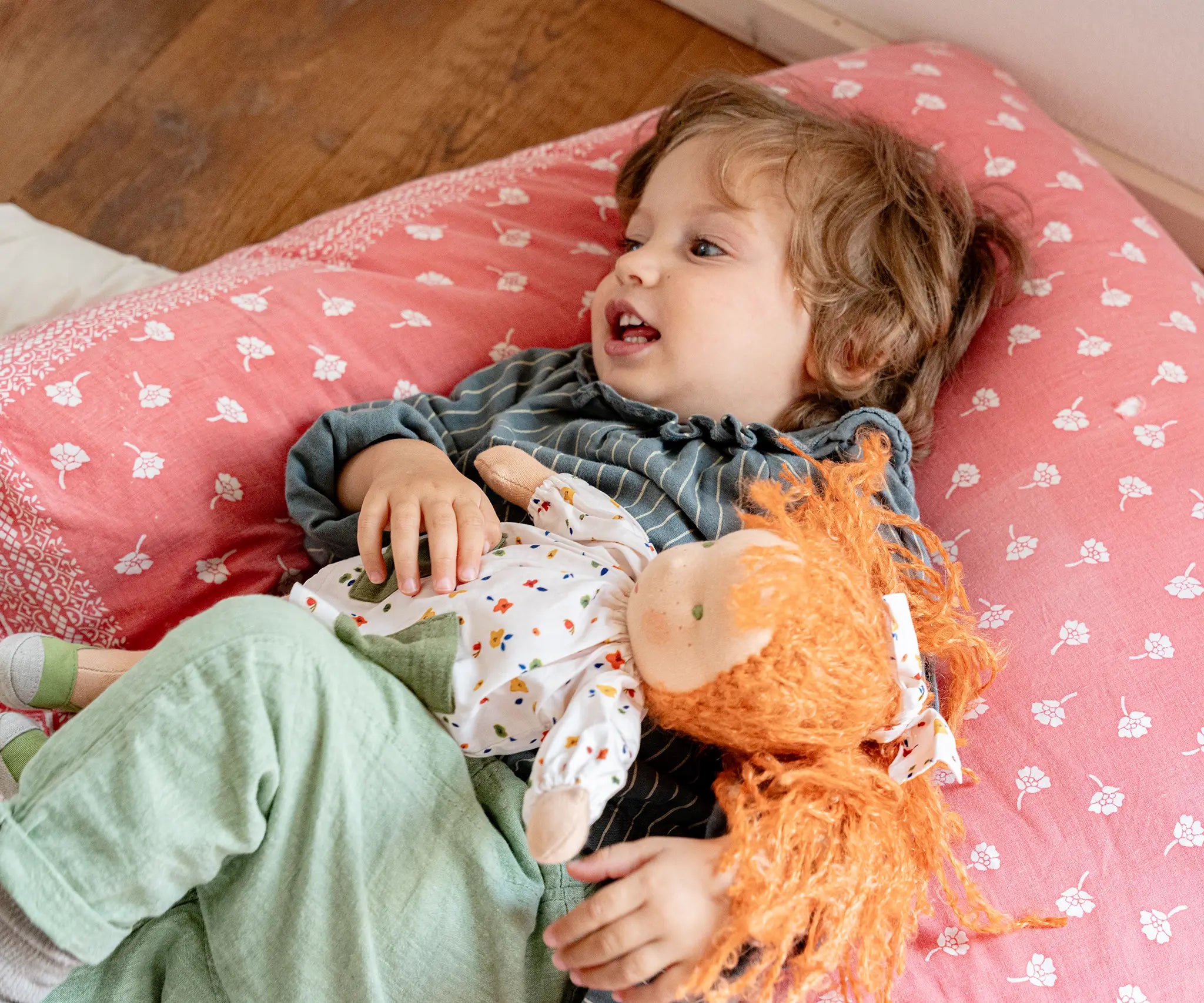 Child laying on a large pink pillow with aKäthe Kruse Waldorf doll Elsa with long red wavy hair and fair skin. White patterned tunic top with pink linen style trousers underneath. White patterned bow in her hair with green and white sneaker style shoes.