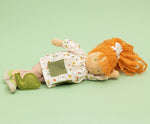 Käthe Kruse Waldorf doll Elsa with long red wavy hair and fair skin laying on her side next to the green felt snail. White patterned tunic top with pink linen style trousers underneath. White patterned bow in her hair with green and white sneaker style shoes.
