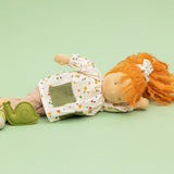 Käthe Kruse Waldorf doll Elsa with long red wavy hair and fair skin laying on her side next to the green felt snail. White patterned tunic top with pink linen style trousers underneath. White patterned bow in her hair with green and white sneaker style shoes.