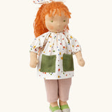 Käthe Kruse Waldorf doll Elsa with long red wavy hair and fair skin. White patterned tunic top with pink linen style trousers underneath. White patterned bow in her hair with green and white sneaker style shoes.