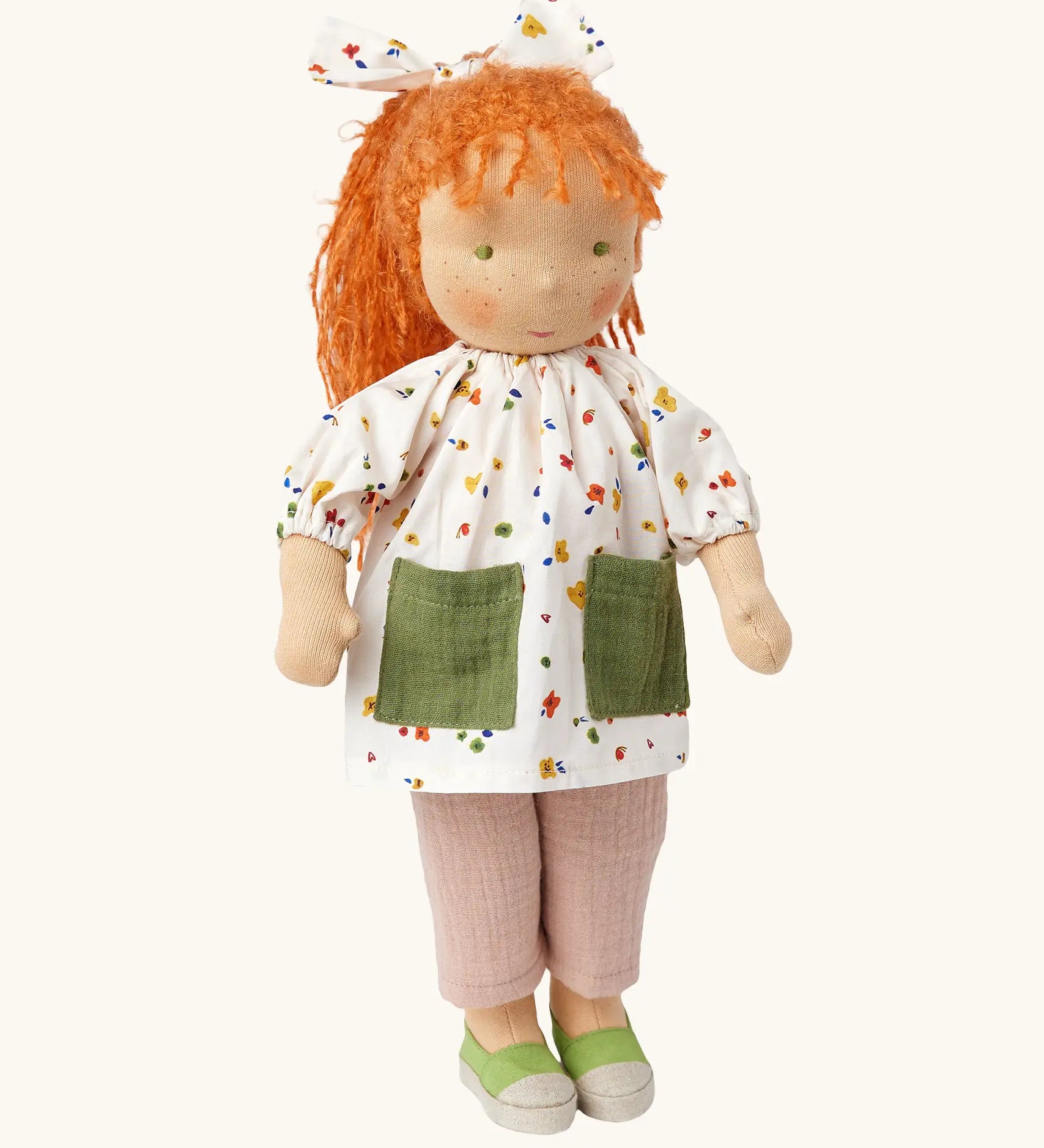 Käthe Kruse Waldorf doll Elsa with long red wavy hair and fair skin. White patterned tunic top with pink linen style trousers underneath. White patterned bow in her hair with green and white sneaker style shoes.