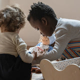 Children playing with a Käthe Kruse Waldorf doll Tilda with long brown hair and fair skin in the background. Blue dotty dress wit red and white striped leggings underneath. White patterned apron and light blue gingham head scarf and red booties.