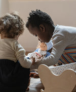 Children playing with a Käthe Kruse Waldorf doll Tilda with long brown hair and fair skin in the background. Blue dotty dress wit red and white striped leggings underneath. White patterned apron and light blue gingham head scarf and red booties.