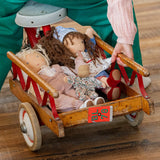 Käthe Kruse Waldorf dolls placed in a wooden trike trailer. Tilda Waldorf doll  with long brown hair and fair skin on the right. Blue dotty dress wit red and white striped leggings underneath. White patterned apron and light blue gingham head scarf and red booties.