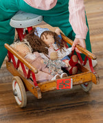 Käthe Kruse Waldorf dolls placed in a wooden trike trailer. Tilda Waldorf doll  with long brown hair and fair skin on the right. Blue dotty dress wit red and white striped leggings underneath. White patterned apron and light blue gingham head scarf and red booties.