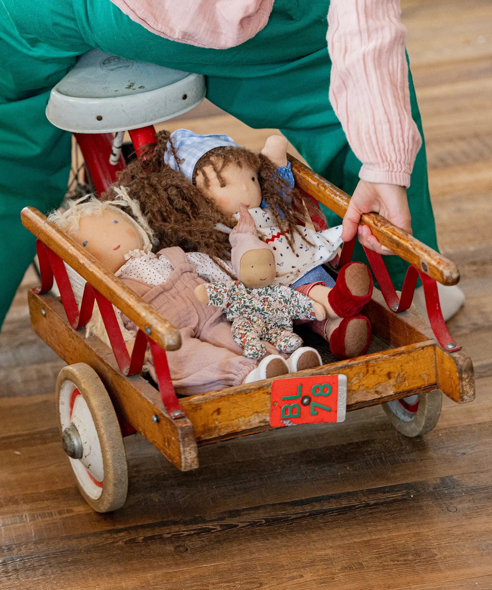 Käthe Kruse Waldorf dolls placed in a wooden trike trailer. Tilda Waldorf doll  with long brown hair and fair skin on the right. Blue dotty dress wit red and white striped leggings underneath. White patterned apron and light blue gingham head scarf and red booties.