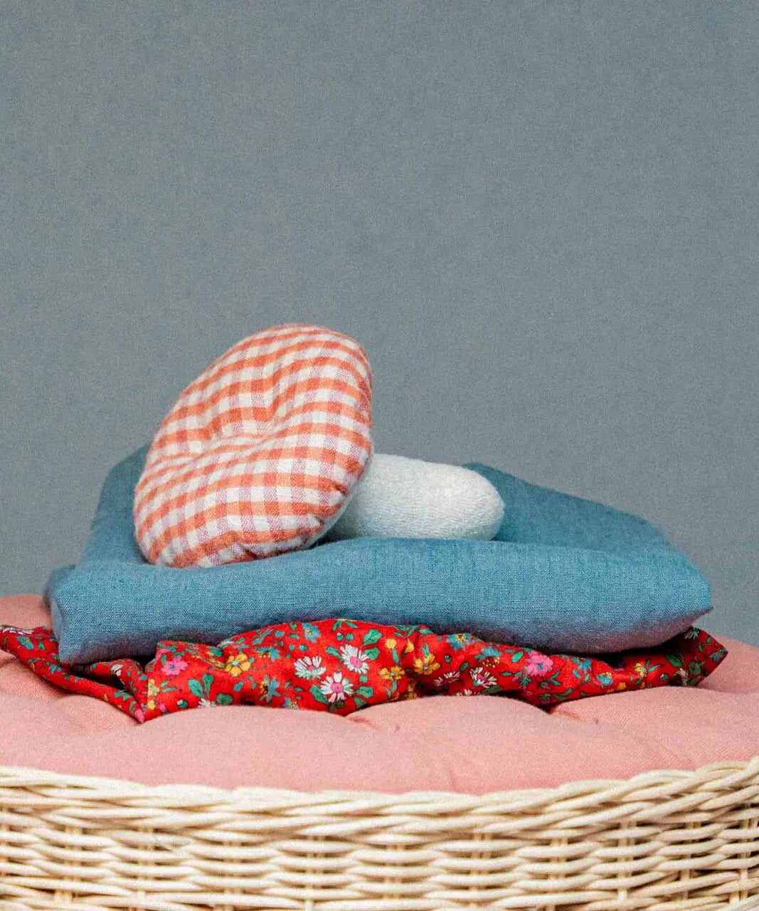 Kathe Kruse mushroom soft grabber toy lying on top of a sheets in a wicker basket. Showing a red gingham top and white stem