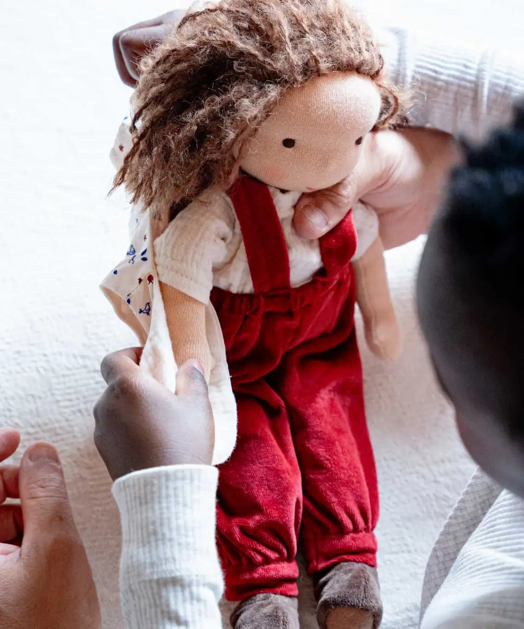 Toni Waldorf Doll by Kathe Kruse, a handmade organic Steiner Doll with red overalls, curly brown hair, and minimal facial features, held by a child. 