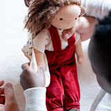 Toni Waldorf Doll by Kathe Kruse, a handmade organic Steiner Doll with red overalls, curly brown hair, and minimal facial features, held by a child. 