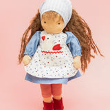 Käthe Kruse Waldorf doll Tilda with long brown hair and fair skin. Blue dotty dress with red and white striped leggings underneath. White patterned apron and light blue gingham head scarf and red booties.