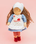 Käthe Kruse Waldorf doll Tilda with long brown hair and fair skin. Blue dotty dress with red and white striped leggings underneath. White patterned apron and light blue gingham head scarf and red booties.