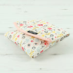 Side view of the Keep Leaf food wrap in bloom design on a wooden surface. A cream colour organic cotton wrap for food with cute flowers printed all over and light pink trim. Features a velcro lid for easy fastening