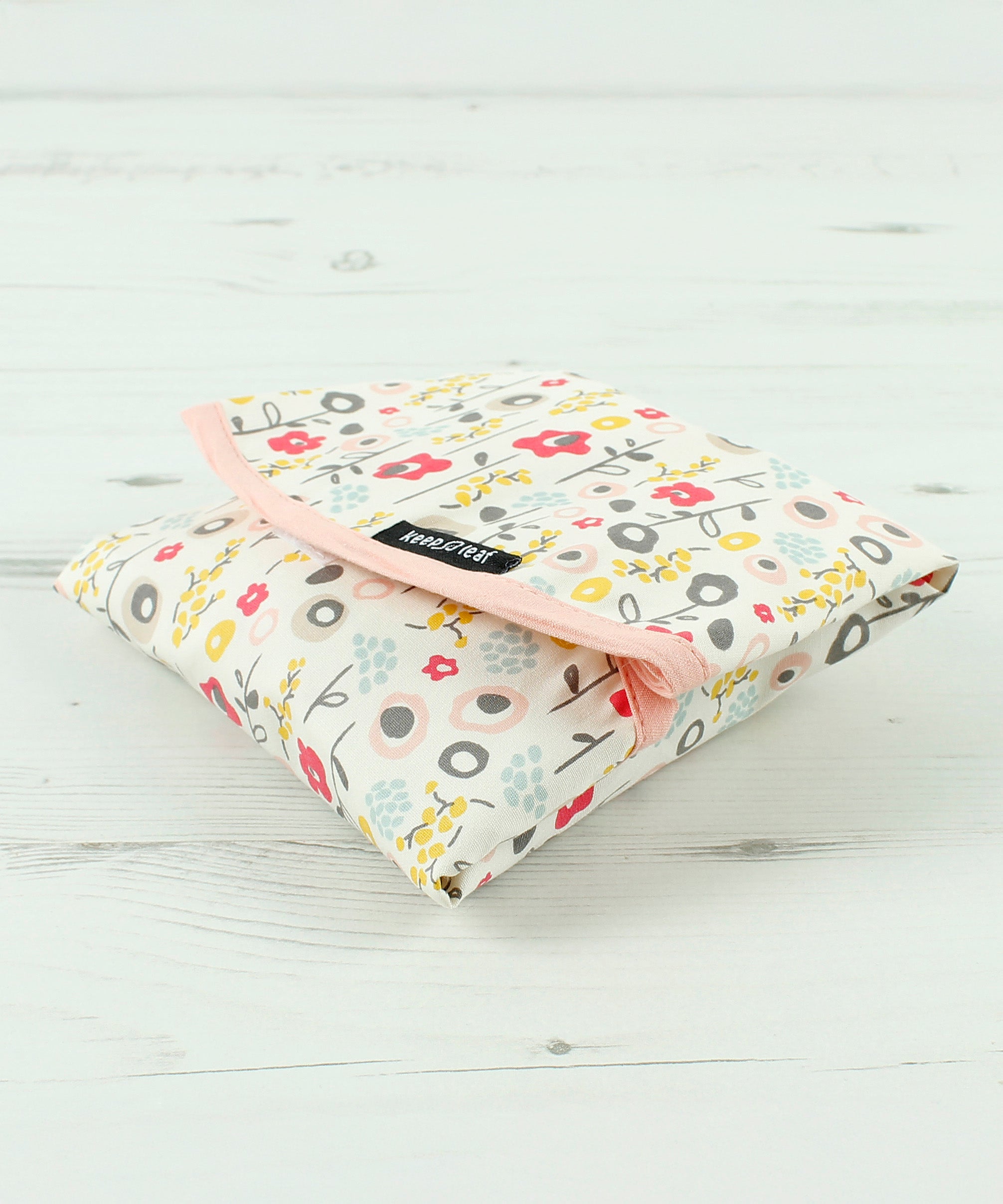 Side view of the Keep Leaf food wrap in bloom design on a wooden surface. A cream colour organic cotton wrap for food with cute flowers printed all over and light pink trim. Features a velcro lid for easy fastening