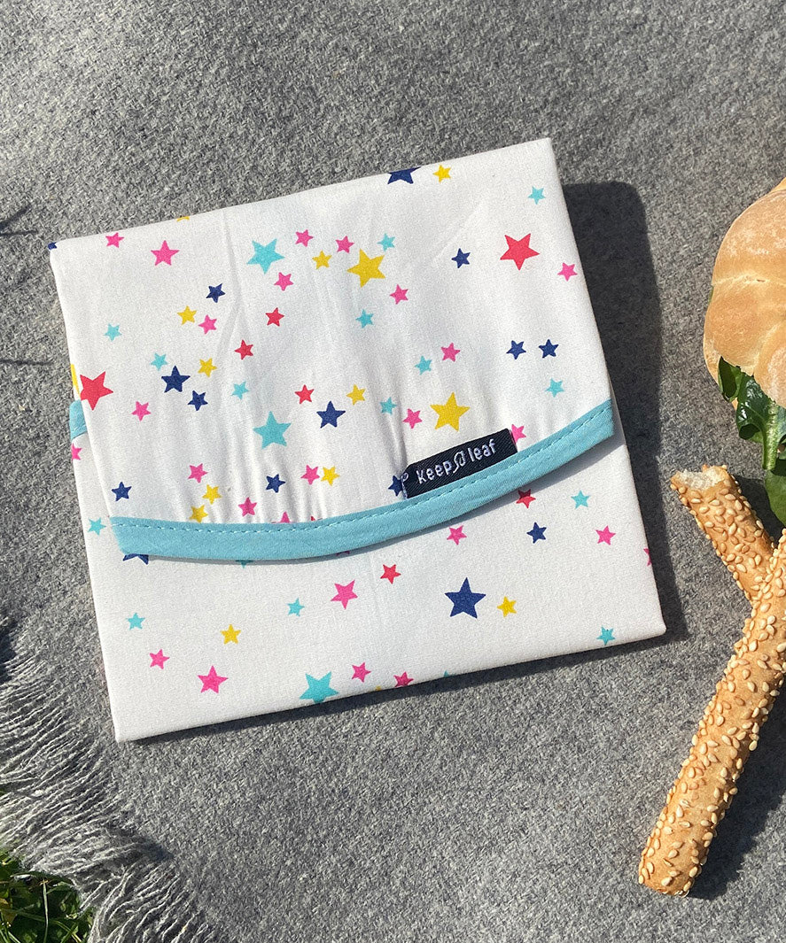 A close up of the Keep Leaf food wrap in stars design on a grey blanket. A cream colour organic cotton wrap for food with multicoloured stars printed all over and light blue trim. Features a velcro lid for easy fastening