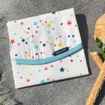 A close up of the Keep Leaf food wrap in stars design on a grey blanket. A cream colour organic cotton wrap for food with multicoloured stars printed all over and light blue trim. Features a velcro lid for easy fastening