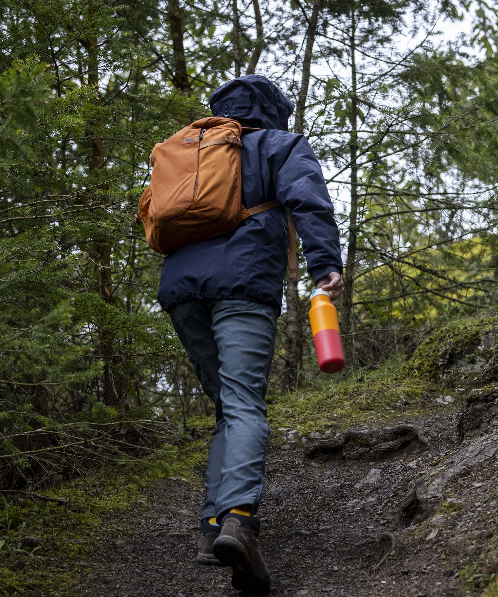 A man walking in woodland and holding the marigold kaleidoscope Klean Kanteen 16oz reusable rise insulated bottle by the white flip sports cap