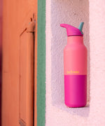 A pink lemonade 16oz Klean Kanteen stainless steel insulated rise bottle standing  on a  white wall 